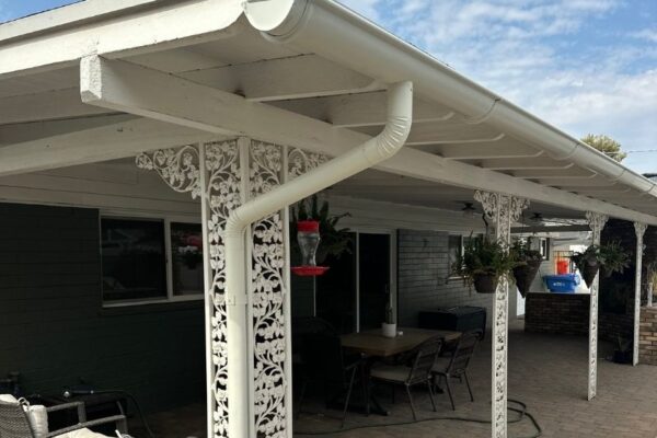 White patio with decorative columns, a table, chairs, and hanging plants under a cloudy sky.