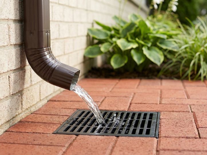 Downspout directing rainwater into a drain on a brick patio, surrounded by lush greenery. Effective drainage solution.