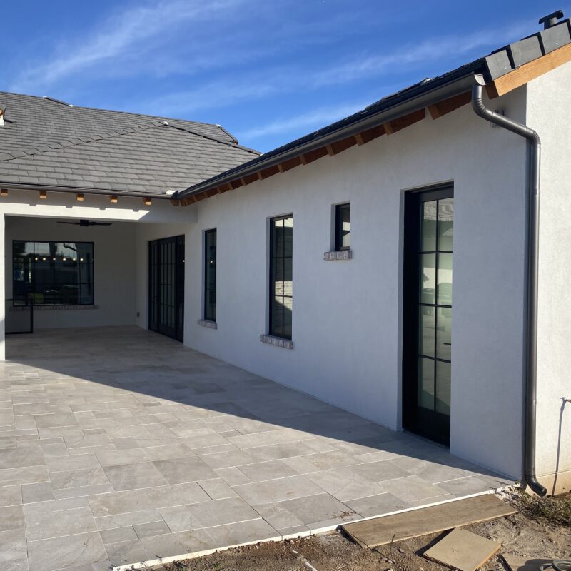 Modern home exterior showcasing a spacious patio with stone tiles and large windows under a blue sky. Perfect for outdoor living.
