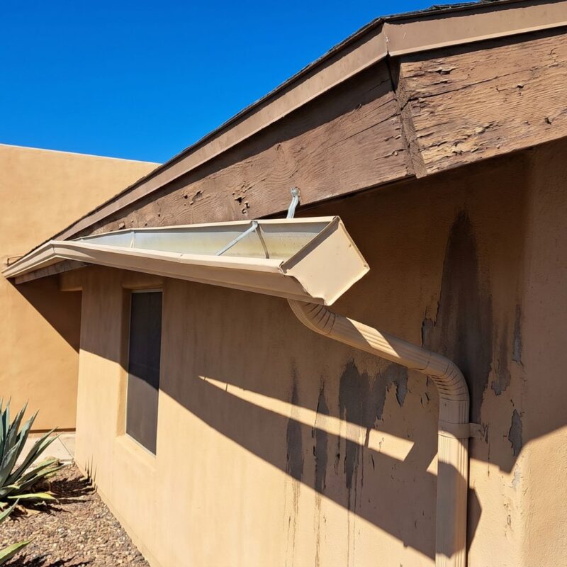 Close-up of a home's exterior showing wear on fascia and gutter, with desert landscaping and clear blue sky.