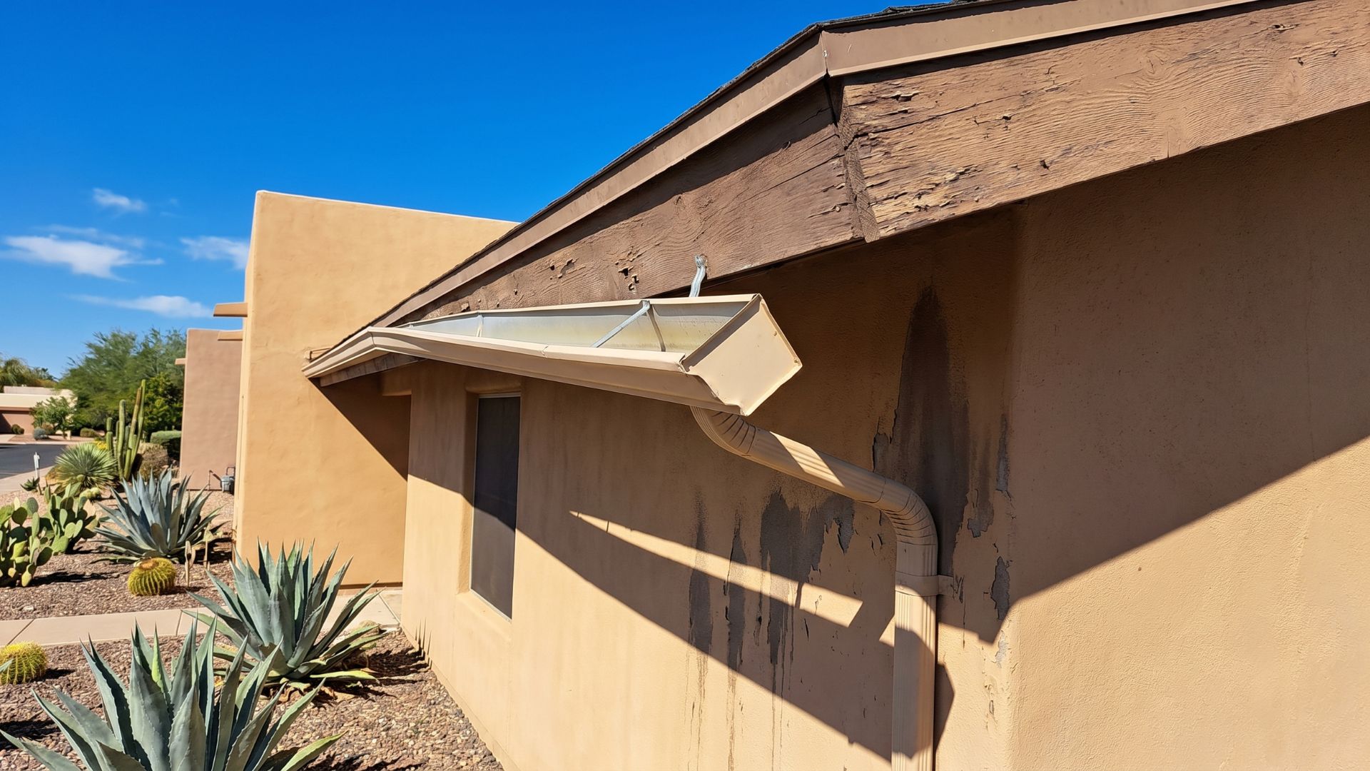 Close-up of a home's exterior showing wear on fascia and gutter, with desert landscaping and clear blue sky.