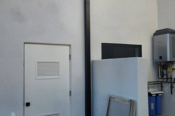 Black vent pipe on exterior wall near a door, with utility equipment and construction materials in view.